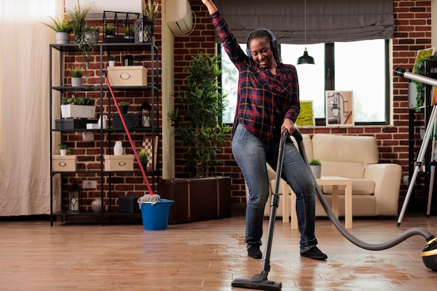 Authentic woman using modern vacuum cleaner listening to favorite music with wireless headphones. Housewife having fun while finishing spring cleaning at home, good vibes and positive energy.