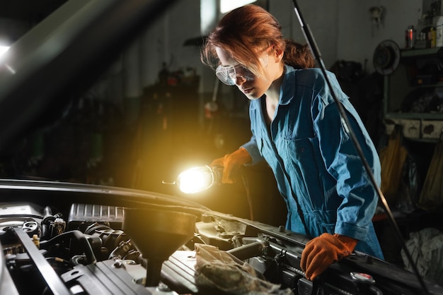 Auto locksmith girl inspects the engine of the car illuminating the light of the lamp Garage or auto repair shop and a woman at work in overalls and glasses