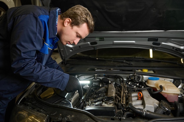 Auto mechanic, technician, car engineer with a flashlight lamp doing a thorough inspection for flaws in the car engine at car repair shop