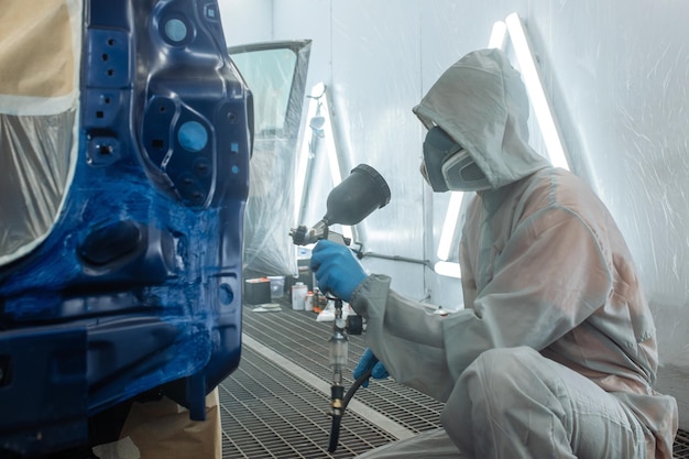 Photo automobile repairman painter hand in protective glove with airbrush pulverizer painting car body in paint chamber.