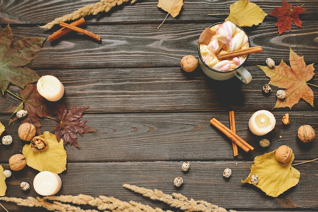 Autumn background made of dried fall leaves, mug of cocoa with marshmallows, nuts, cinnamon, plaid, apples. Top view on brown wood