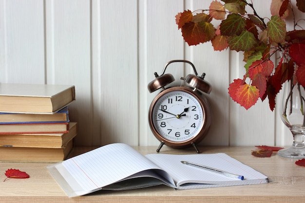 Autumn composition with an open notebook, textbooks and autumn aspen leaves on the table back to school education study interior with a bronze alarm clock