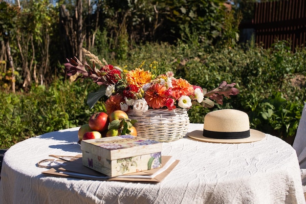 An autumn flower arrangement in a basket is on the table next to a hat and apples