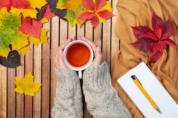 Autumn leaves, white notebook, pen, tea cup in female hands in knitted sweater, textile napkin, wooden background, mockup, place for text