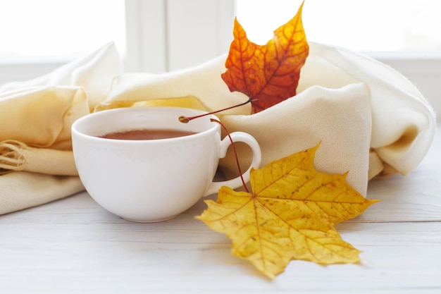 Autumn leaves with a cup of tea or coffee next to a scarf on a wooden background with copy space