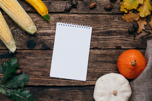 Autumn mood: white notebook, pumpkins and corn with yellow leaves on wood