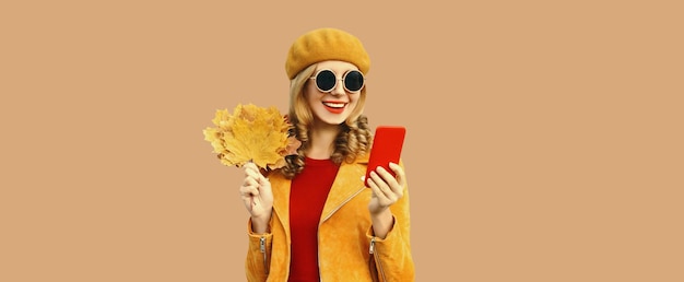 Autumn outfit portrait of fashionable smiling young woman with phone holds yellow maple leaves