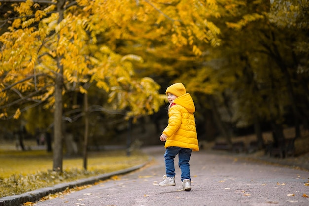 Autumn in the park. Back view of little cute girl in stylish yellow coat and cap, running, jumping and having fun, while walking in beautiful golden autumn park.