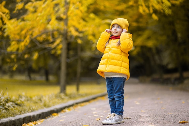 Autumn in the park. Cute dreamy little girl in a yellow cap and warm clothes standing on a park alley with golden trees background and looking up. Smiling kid walking in autumn park