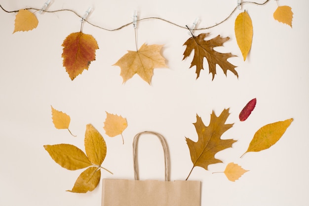 Autumn shopping with discounts. Autumn sales. Kraft beige paper shopping bag, from which look out autumn yellow leaves. Copy space. On a thread hanging on clothespins autumn leaves