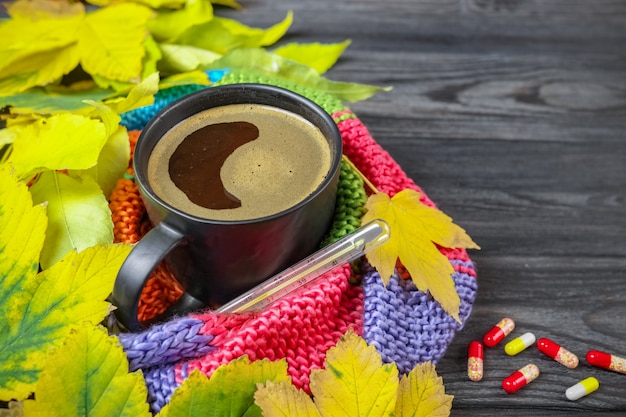 Autumn still life: A cup of hot coffee and a thermometer wrapped in a warm scarf, yellow leaves.