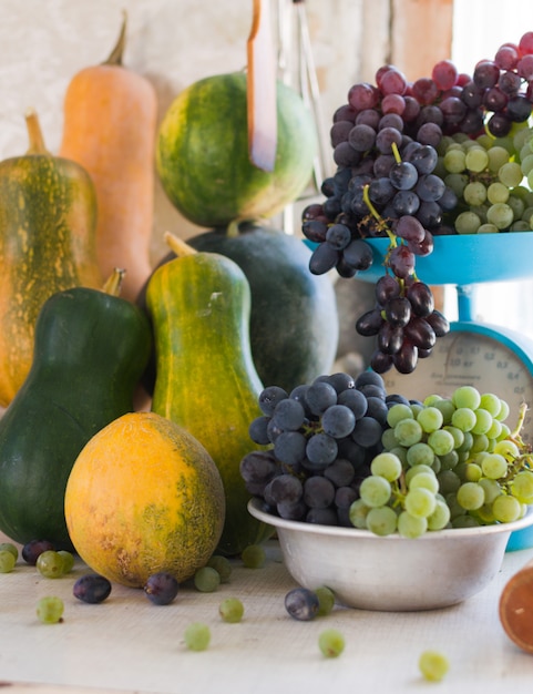 Autumn still life with pumpkins, melons, watermelon, grapes on a scale and in a metal bowl on a wooden white table. Autumn harvest concept.