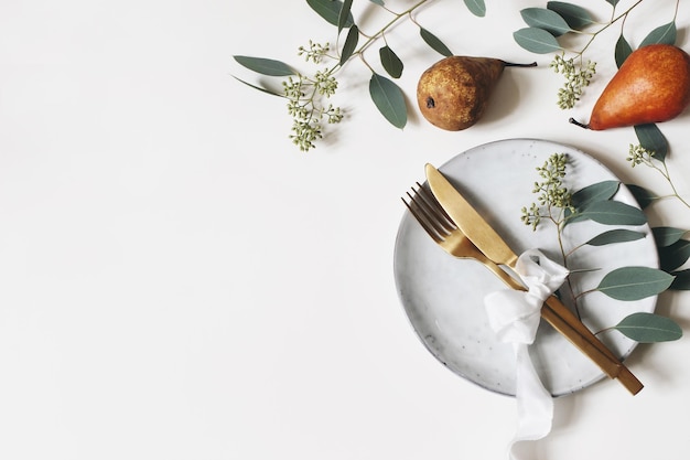 Autumn thanksgiving table place setting Golden cutlery porcelain plate berry eucalyptus leaves and branches silk ribbon and pear fruit on white table background Fall wedding Flat lay top view