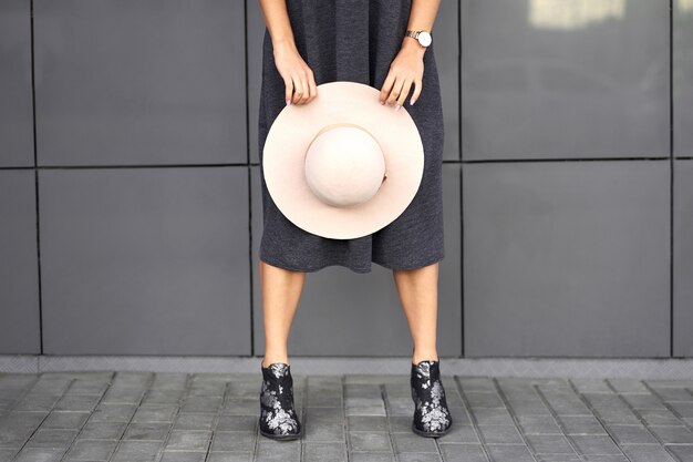 Autumn trendy collection. Happy beautiful brunette girl wearing stylish grey dress holding fashionable hat in the hands. Model in trendy outfit posing on grey wall background. Outdoor portrait.