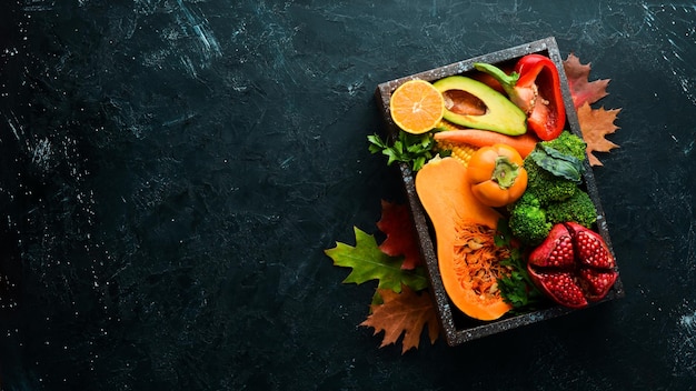 Autumn vegetables and fruits on a black stone background: Pumpkin, tomatoes, corn, pomegranate, persimmon, apple. Top view. Free copy space.