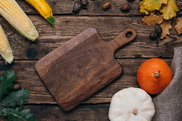 Autumn vegetables: pumpkins and corn with yellow leaves and cut board on wood