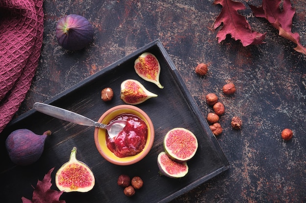 Autumntime background with fig jam spoon nuts and fresh halved fig fruits on black wooden tray Dark brown background with dry Fall leaves