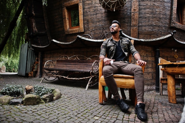 Awesome beautiful tall ararbian beard macho man in glasses and military jacket posed outdoor sitting on chair against wooden house