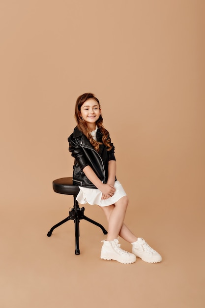 Awesome girl. Full length of a fun little girl, cross-legged and looking with a smile, sitting on a chair on a beige wall.