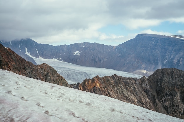 Awesome mountain scenery with layers of rocks and mountains with snow and glacier in high altitude under cloudy sky. Scenic view from snow cornice to glacier tongue among sharp rocks in low clouds.