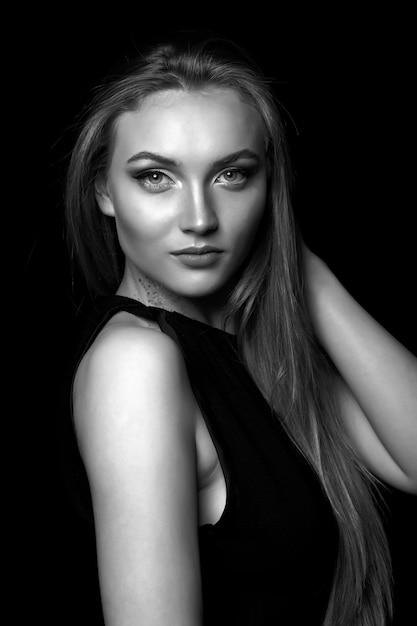 Awesome young woman with long straight hair wears classic black dress posing in the shadow at studio. Black and white shot