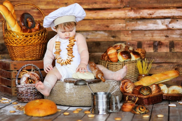 A baby baker boy in a chef's hat and apron smeared in flour with a bunch of bagels around his neck prepares dough for baking.