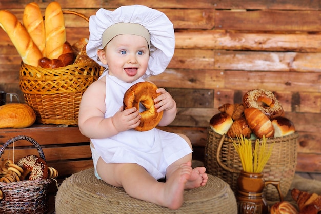 Baby baker boy in a chefs hat and apron with a large bagel in his hands smiles against the background of bakery products