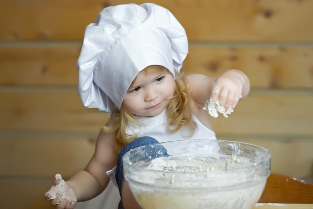 Baby baker or child with happy face in white cook uniform with chef hat and apron kneading dough with flour in glass bowl in kitchen on wooden 