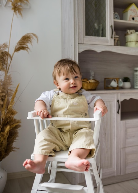 A baby boy in a beige jumpsuit is sitting in a white high chair in the kitchen Child's diet