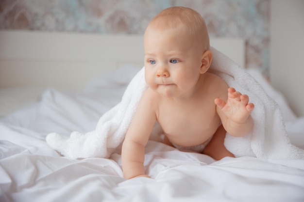 A baby boy in a diaper is sitting on the bed covered with a towel after bathing in the bedroom