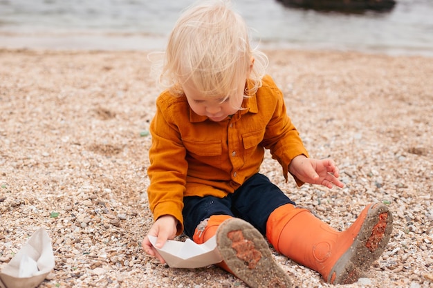 Baby boy is playing with paper boat on the beach near sea Child on vacation in summer or autumn at the sea autumn or summer time