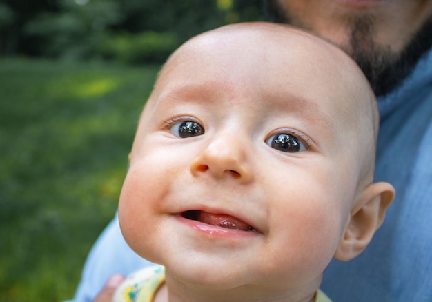 Baby boy looking at the camera and smiling close-up. Dad holds his newborn son in his arms outdoors.