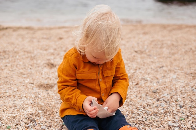 Baby boy playing with paper boat in the sand in autumn time