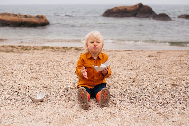 Baby boy playing with paper boat on sand on beach in autumn time