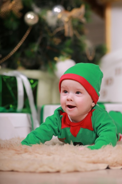 Baby boy in red green elf costume sitting under Christmas tree and gift boxes.