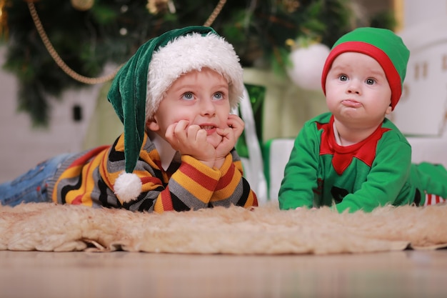 Baby boy in red green elf costume with his older brother in santa hat sitting under Christmas tree and gift boxes.