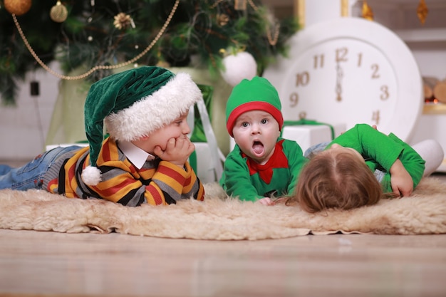 Baby boy in red green elf costume with his older brother and sister in santa hats sitting under Christmas tree with gift boxes.