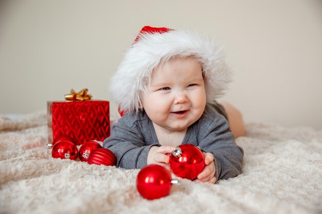 A baby boy in a Santa Claus hat is lying on the bed playing with Christmas balls