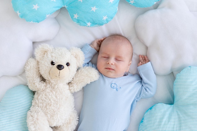 Baby boy sleeping on the bed lying on his back with a soft teddy bear among the pillows in blue pajamas healthy newborn sleep