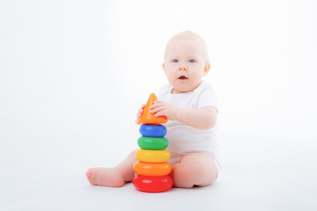 baby boy in white bodysuit is sitting playing with multicolored pyramid smiling on white background