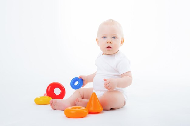 baby boy in white bodysuit is sitting playing with multicolored pyramid smiling on white background