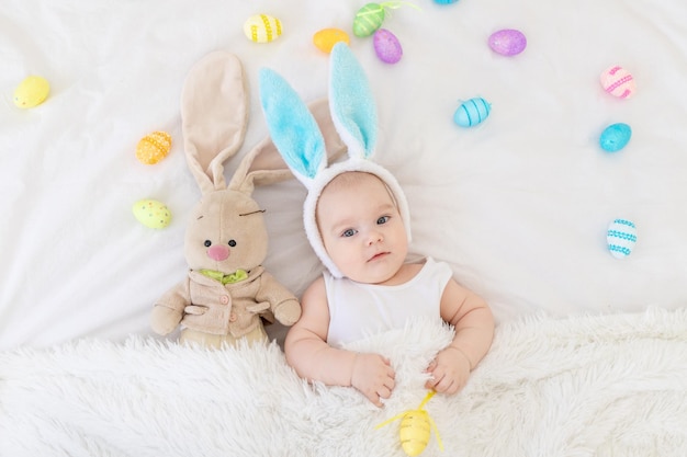 A baby boy with rabbit ears on his head is lying in a crib with a bunny toy and Easter eggs a cute funny smiling little baby Easter Concept