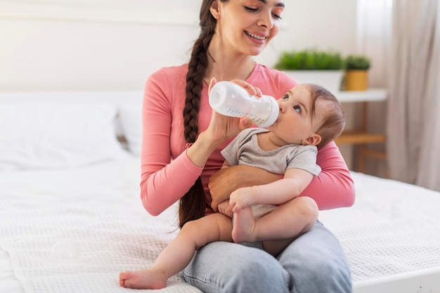 Baby care Loving mom giving bottle with water or milk to her little daughter while relaxing together on bed copy space