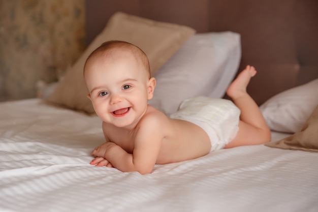 The baby in a diaper is lying on the bed in the bedroom on his stomach smiling