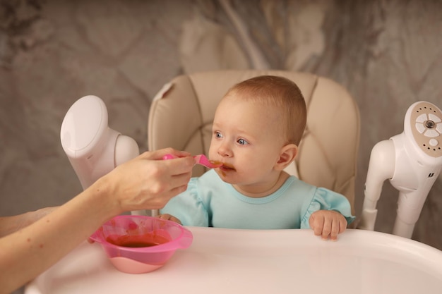 the baby in the feeding chair is fed by her mother with fruit and vegetable puree from a spoon