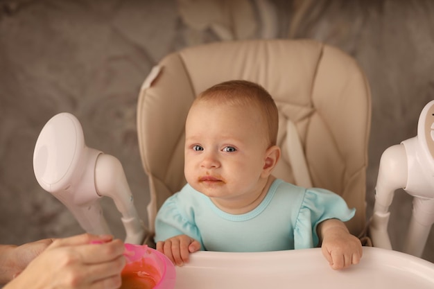 the baby in the feeding chair is fed by her mother with fruit and vegetable puree from a spoon