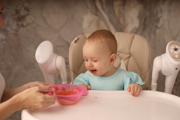 the baby in the feeding chair is fed by her mother with fruit and vegetable puree from a spoon