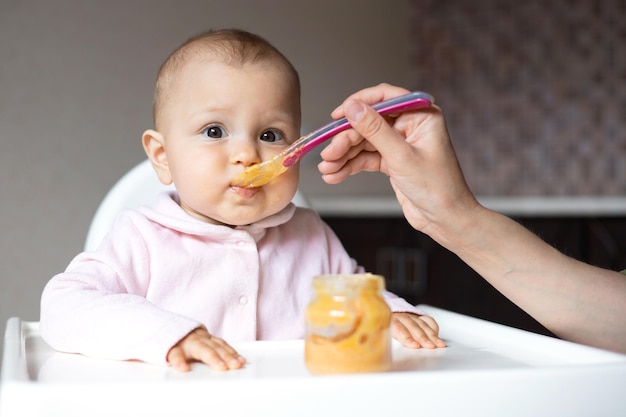 Baby food A baby in a high chair eats vegetable puree from a spoon Mom feeds the baby from a jar Kitchen Lifestyle