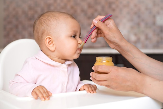 Baby food A mother feeds her baby daughter with a spoonful vegetable puree from a jar The child sits in a high chair in the kitchen