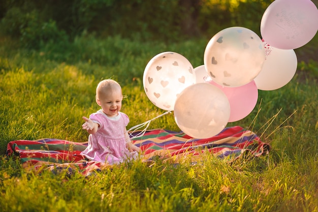 Baby girl 1 year old sitting on green grass with pink and white balloons in meadow outdoors closeup